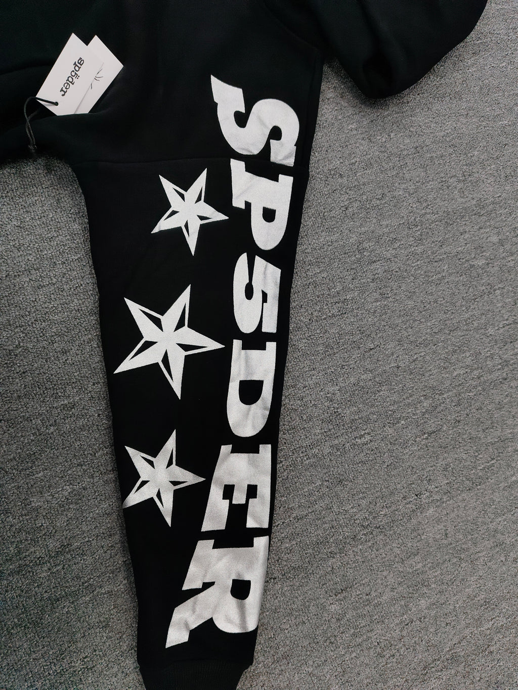a Sp5der MX555 Zip Hoodie in black, featuring white graphic lettering and star prints on the sleeves