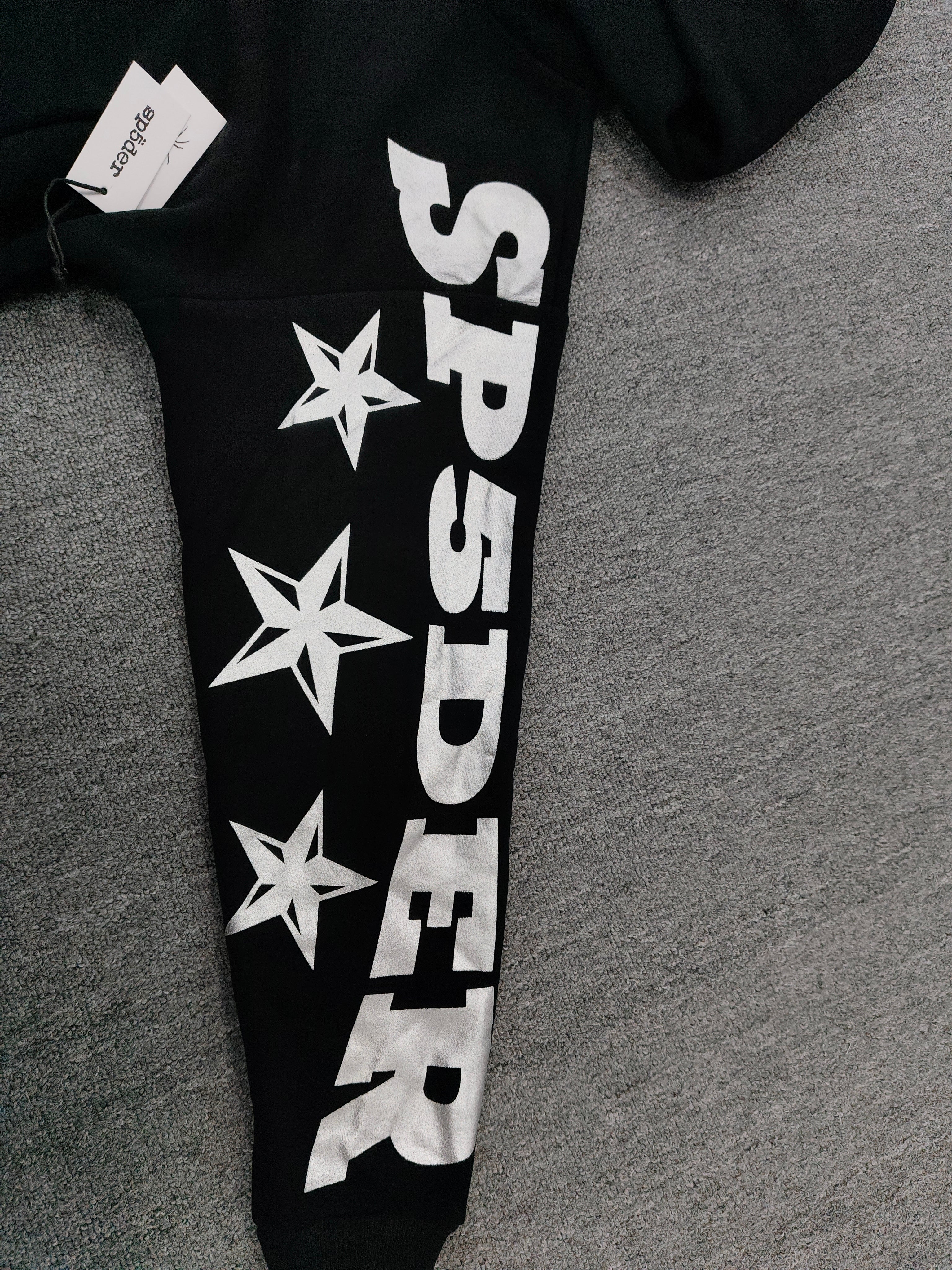 a Sp5der MX555 Zip Hoodie in black, featuring white graphic lettering and star prints on the sleeves