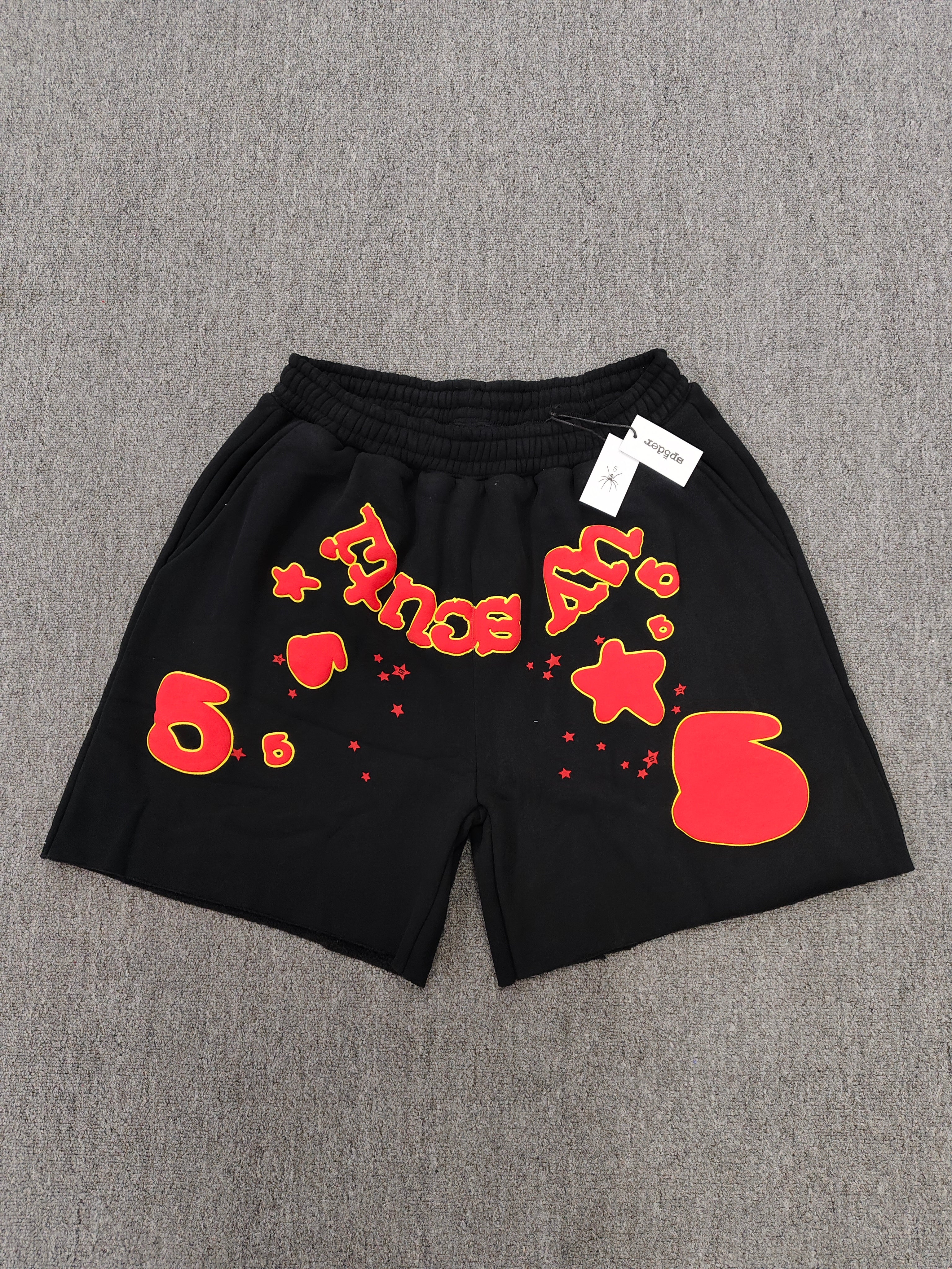 Sp5der x UY SCUTI collaboration shorts featuring bold red graphic logos on a black base