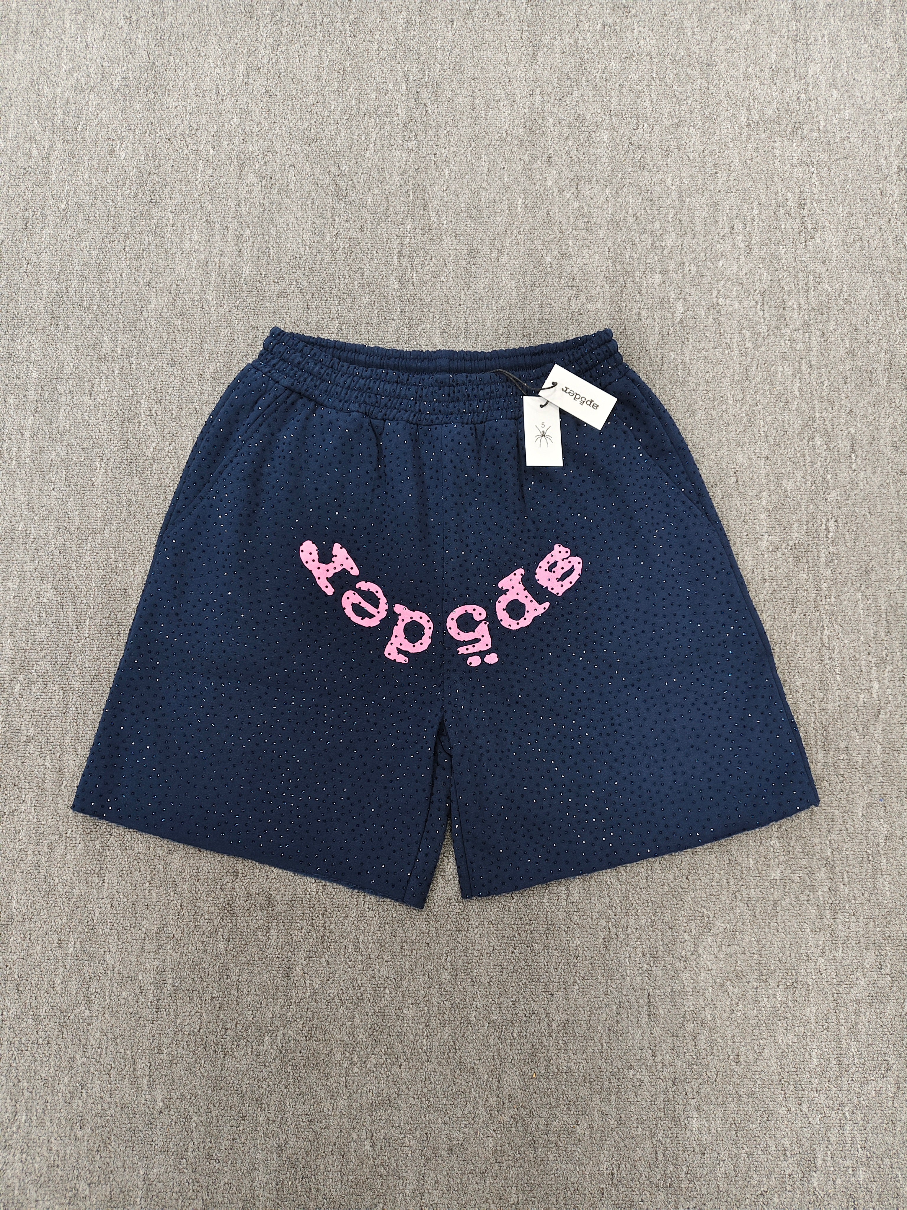 Sp5der jogger shorts are a navy blue color with a pink text graphic and rhinestone details