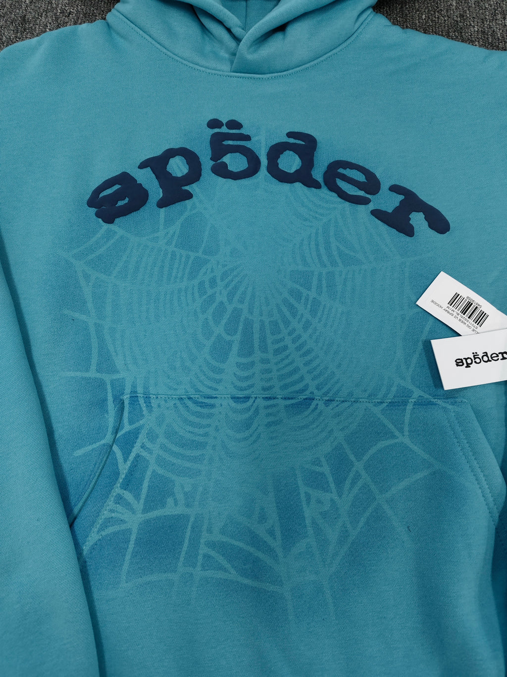 light blue Sp5der OG Web V2 Spray Hoodie features a prominent, sprayed-on navy spiderweb graphic
