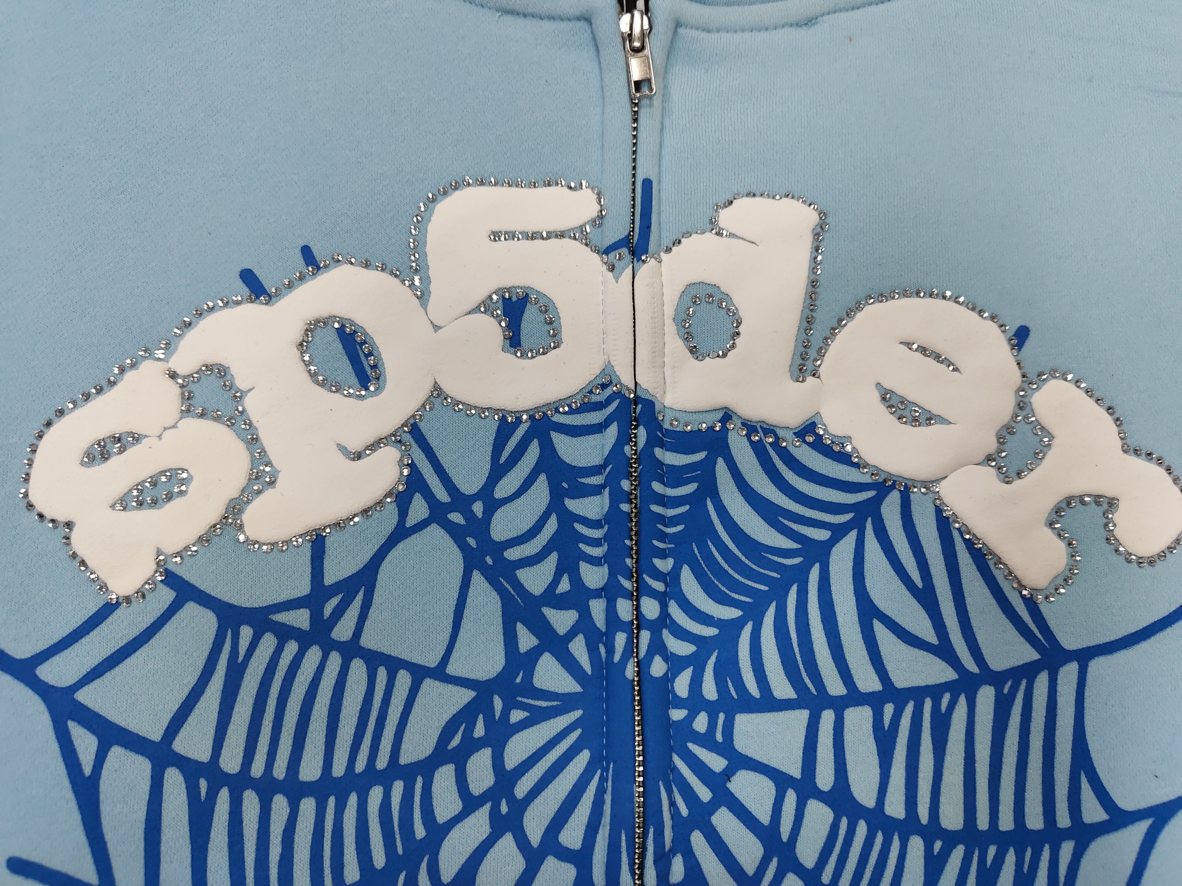 Sp5der OG Web Zip Up hoodie in Sky Blue features a prominent 3D puff-printed spider web graphic and rhinestone embellishments