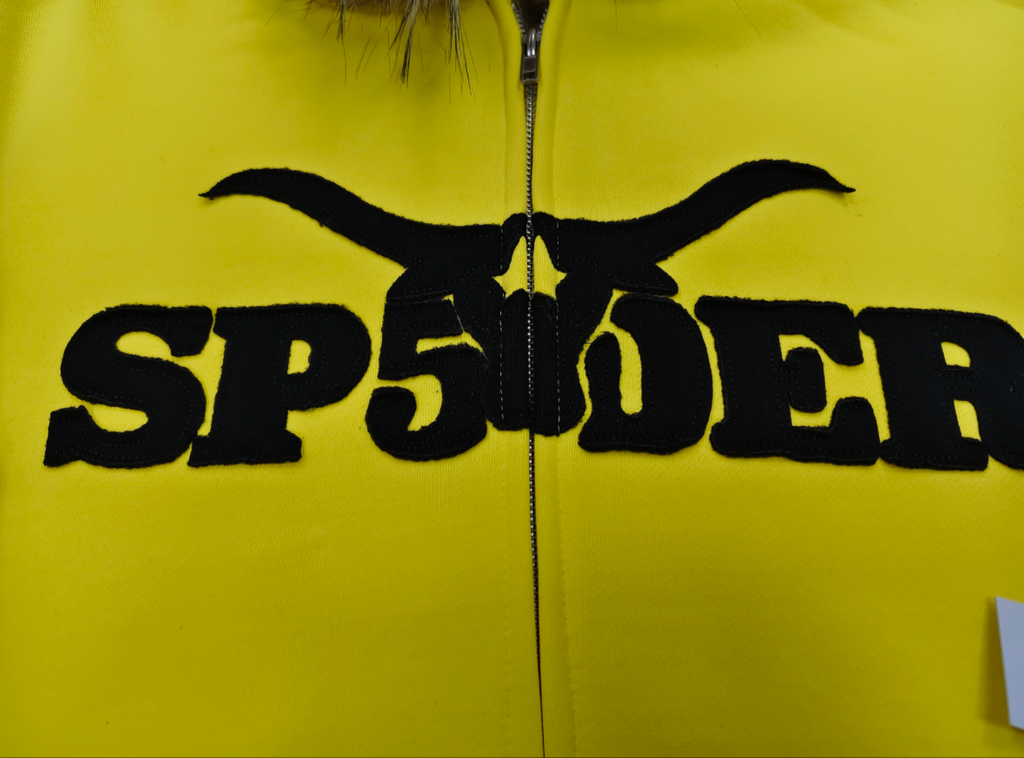 a yellow Sp5der fur-hooded zip-up sweatshirt featuring a prominent black logo and studded sleeve details