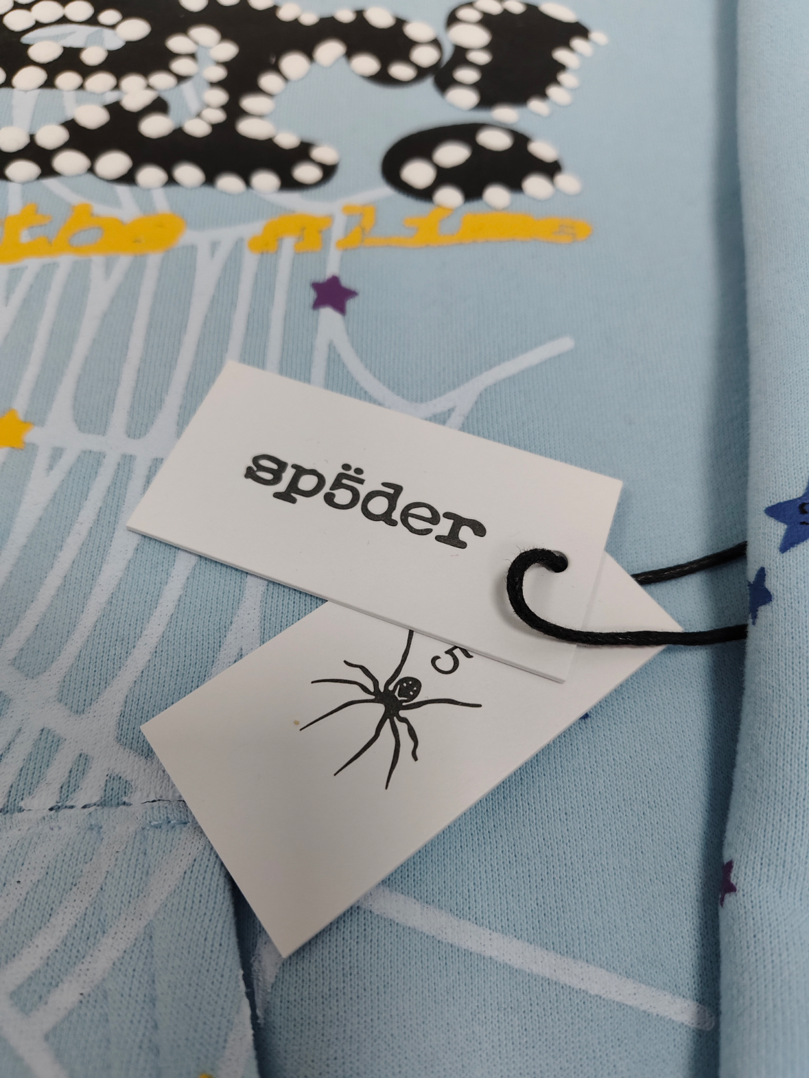 a light blue Sp5der Worldwide hoodie featuring a prominent black graphic logo and a white spiderweb design filled with colorful stars